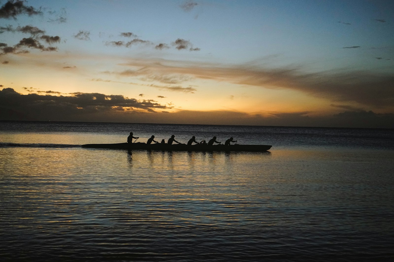 An outrigger canoe crew paddles in silhouette at dusk, low golden light on calm water.